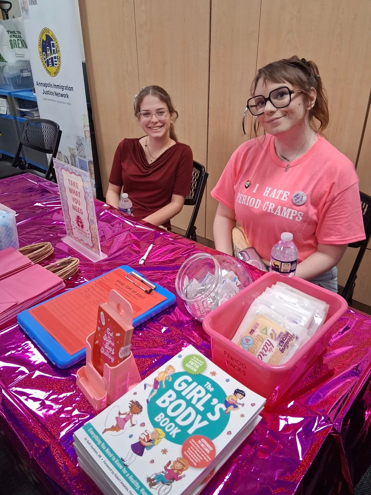Two volunteers seated at a colorful resource table with period supplies and educational materials.